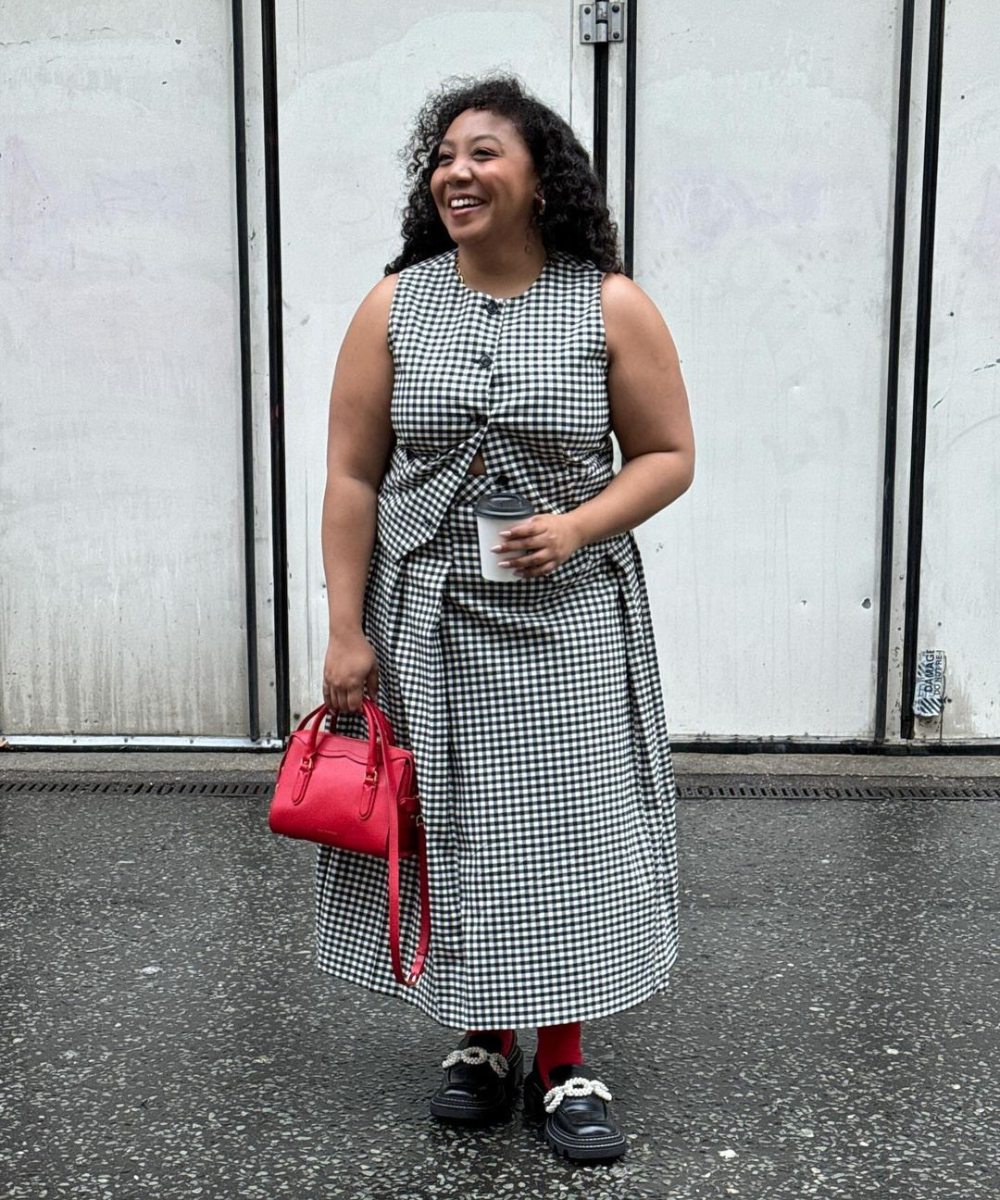 Mulher sorrindo com vestido xadrez vichy, bolsa vermelha e sapatos pretos na calçada.