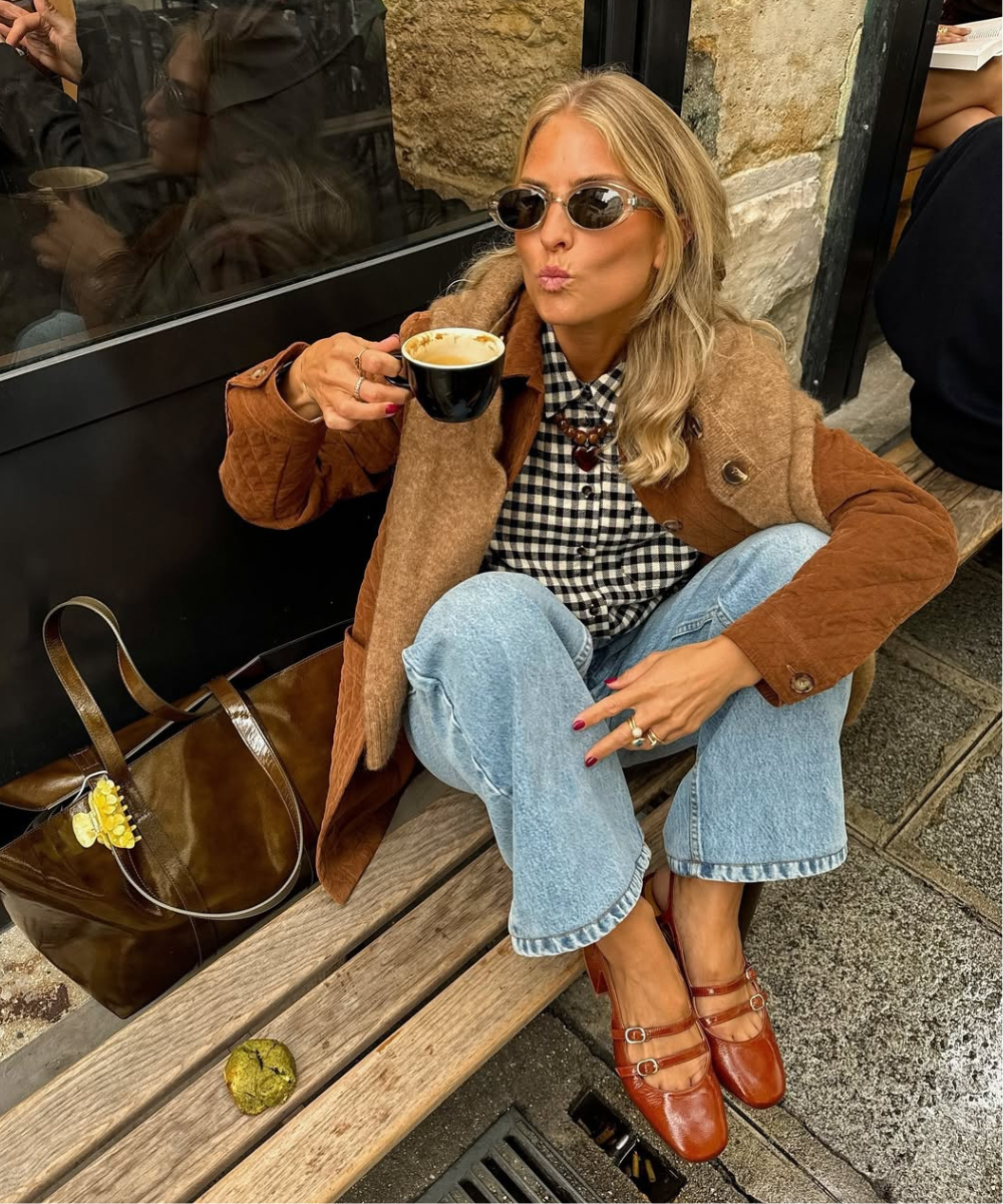 Mulher com óculos escuros em blusa vichy segurando uma xícara de café, usando casaco marrom e calça jeans rasgada.