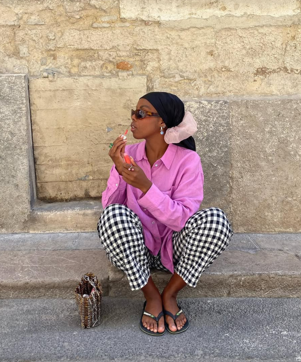 Mulher em blusa rosa se prepara, usando calça xadrez vichy, enquanto sentada em frente a uma parede de pedra.