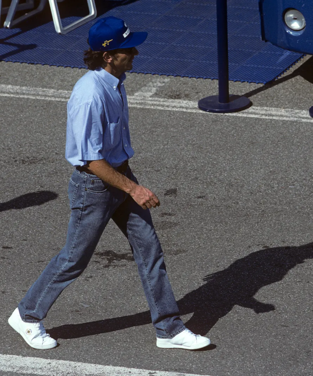 Homem de jeans, tênis branco e boné azul caminhando em pista cinza, estilo de Ayrton Senna.