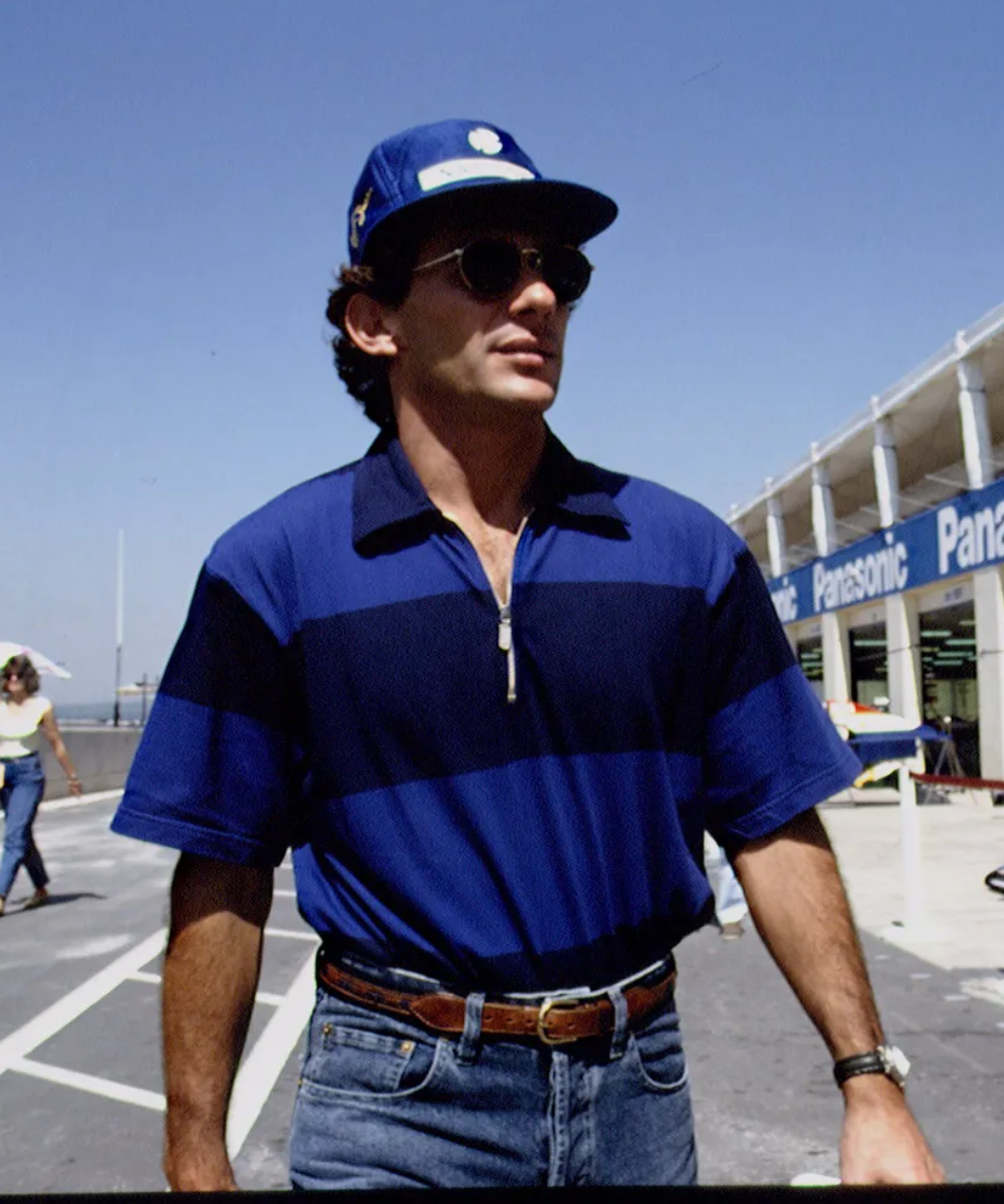 Homem com boné azul, óculos escuros e camisa listrada, estilo descontraído de Ayrton Senna.