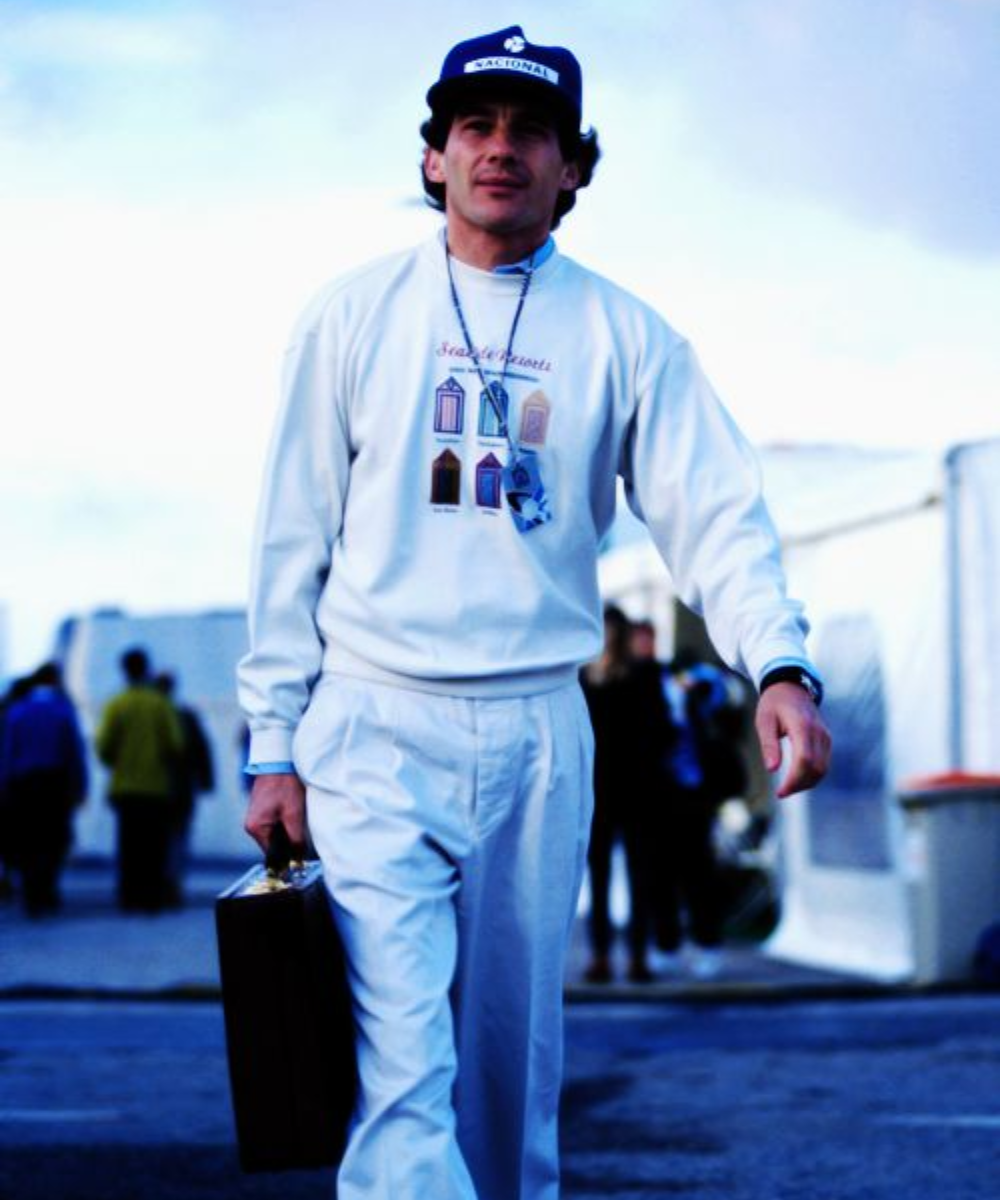 Homem com boné azul e roupa casual branca, carregando uma pasta, remete ao estilo de Ayrton Senna.