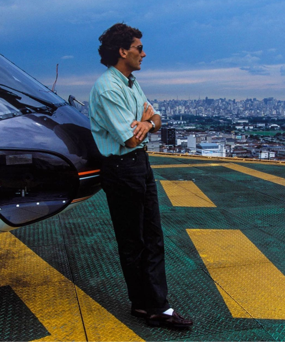 Homem de óculos escuros ao lado de um helicóptero, com vista de cidade ao fundo, em um estilo casual e confiante.