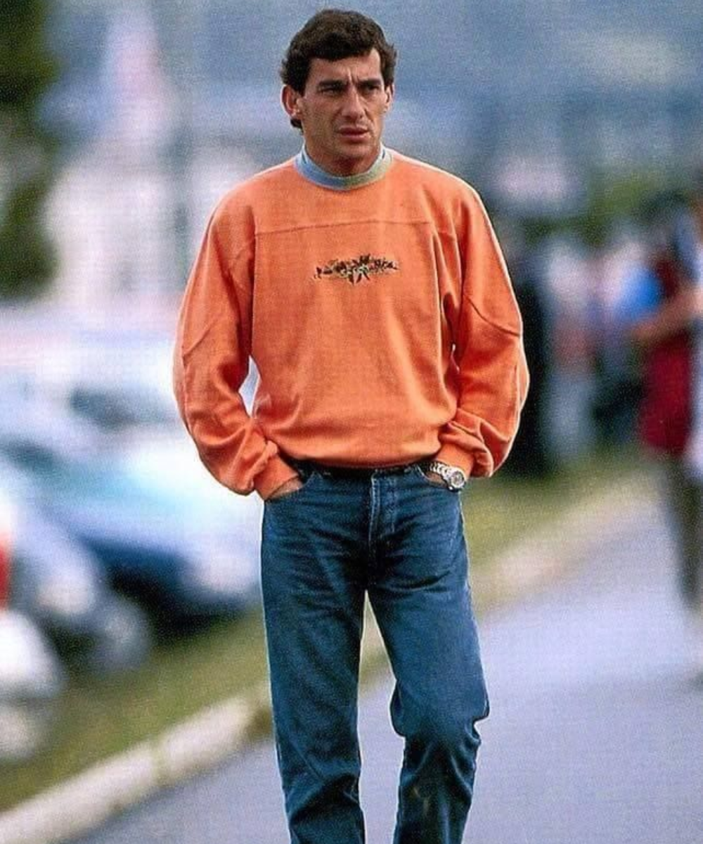 Homem usando suéter laranja e jeans em cenário urbano, caracterizando o estilo sóbrio e confortável dos anos 90.