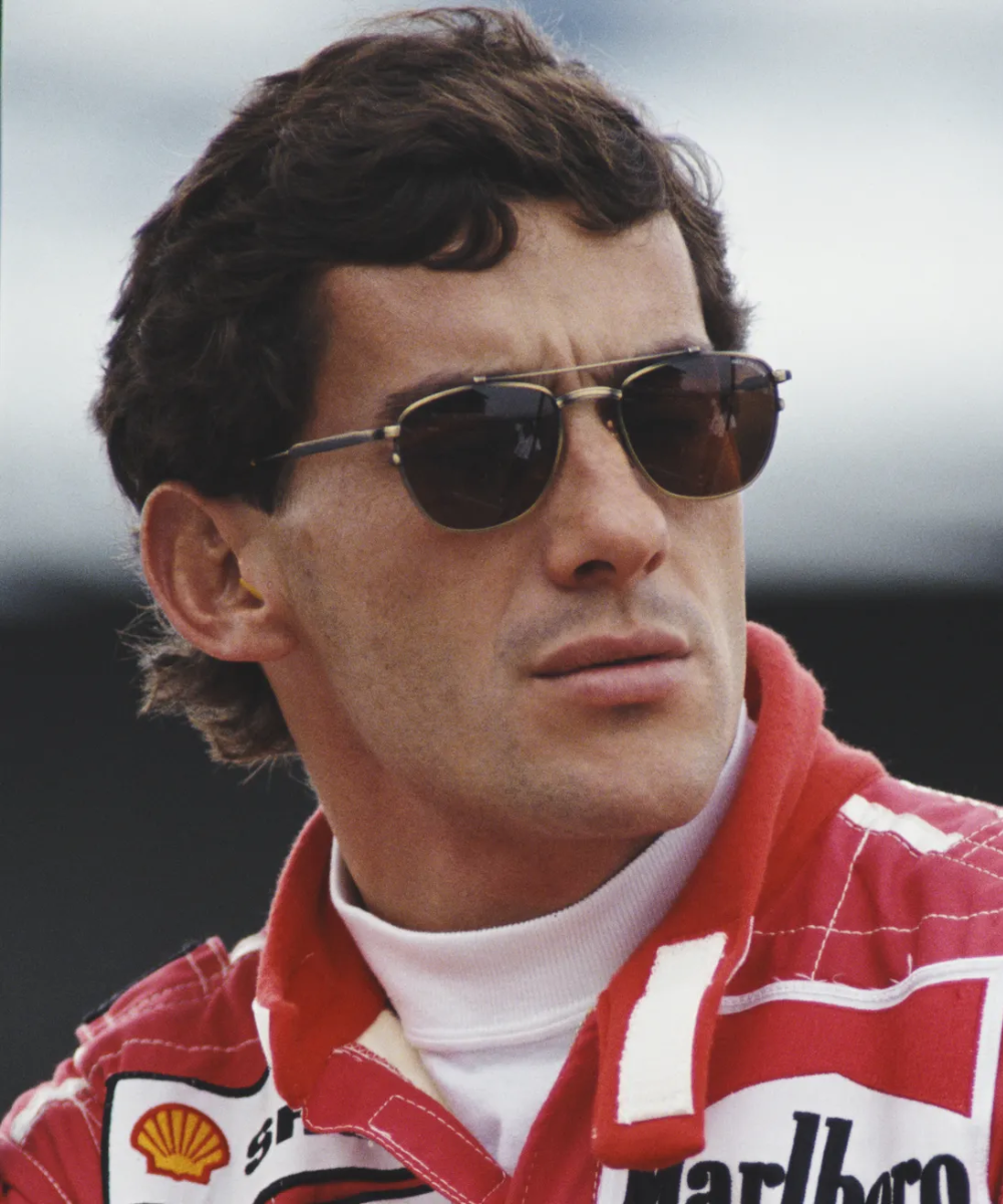 Piloto com estilo icônico de Ayrton Senna, usando óculos escuros e macacão de corrida vermelho.