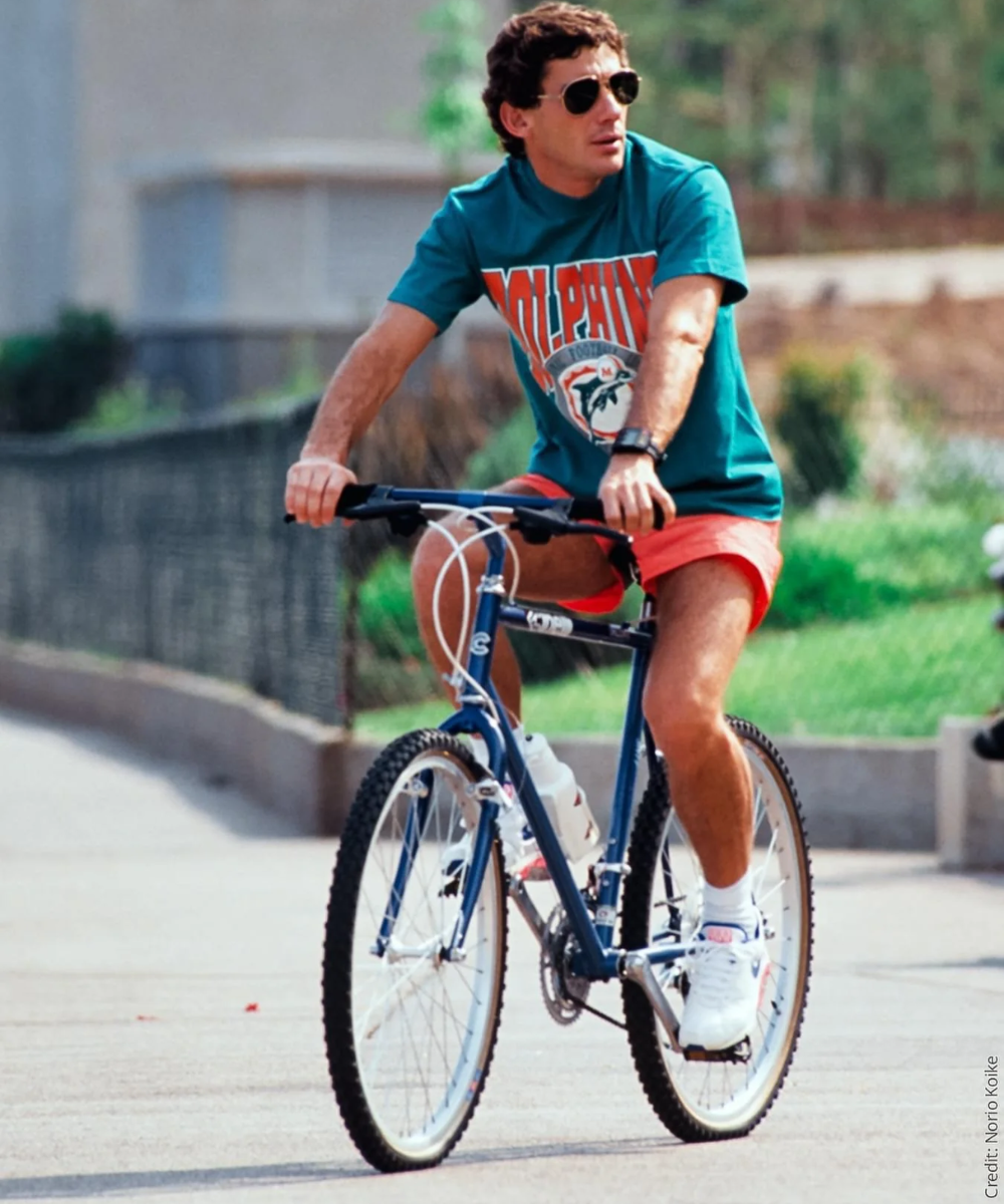 Pessoa de óculos de sol pedalando bicicleta, vestindo camiseta esportiva e shorts. Aparência casual e descontraída.