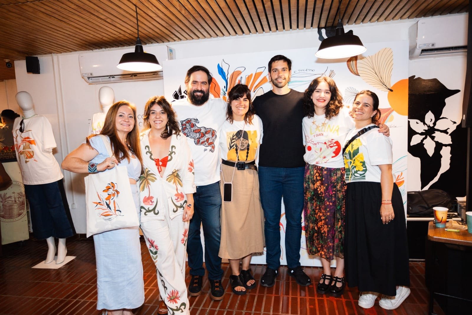 Pessoas sorrindo em evento; algumas vestem roupas coloridas com estampas de palmeiras e ilustrações. A palavra 