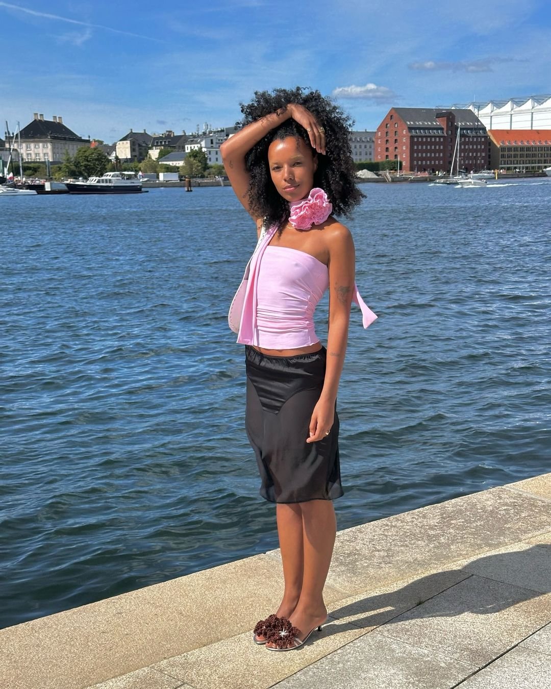 Mulher com vestido rosa e flor, em pé à beira de um rio ensolarado com prédios ao fundo.