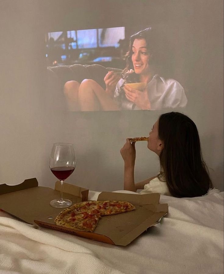 Pessoa assiste a filme de romance projetado na parede enquanto come pizza. Uma taça de vinho está ao lado.