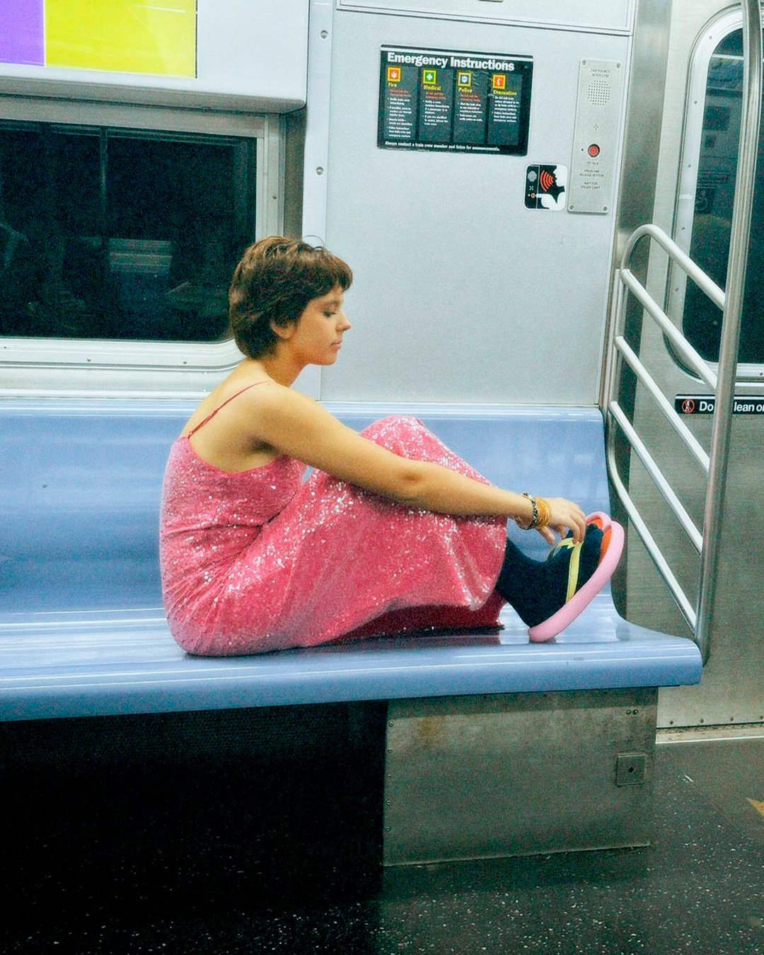 Pessoa sentada no metrô, vestindo um vestido rosa brilhante, calçando chinelos brilhantes.