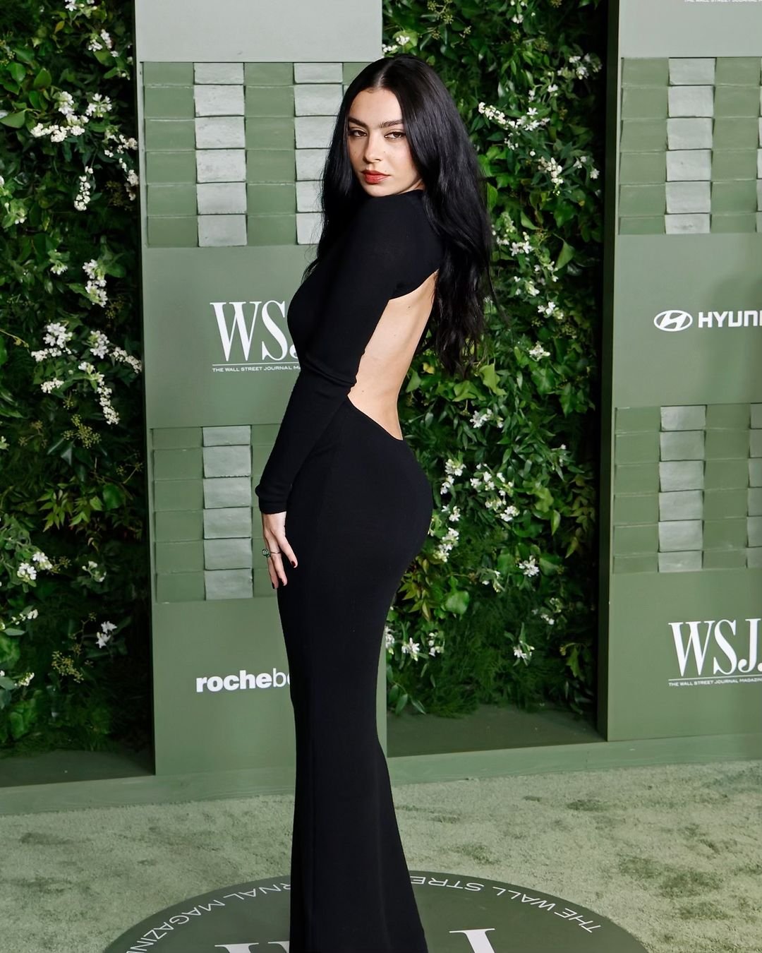 Pessoa com vestido preto elegante em um evento sofisticado, posando em frente a um fundo de folhas verdes. Goth Girl.