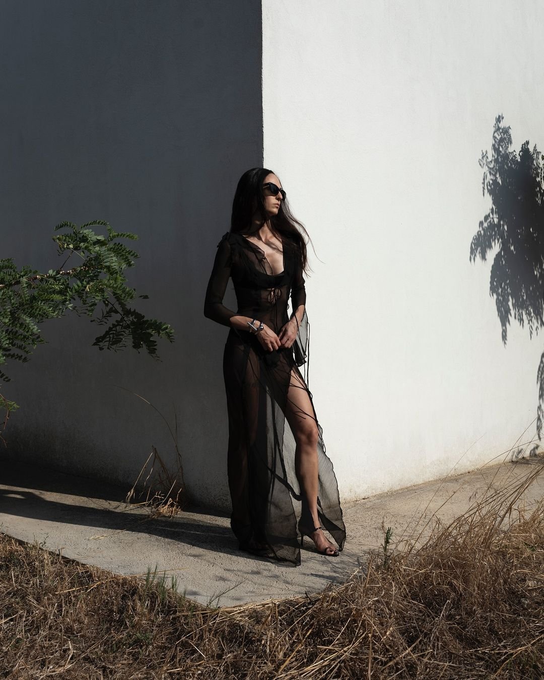 Mulher de vestido preto transparente e óculos escuros em cenário urbano com sombra; estilo goth girl.