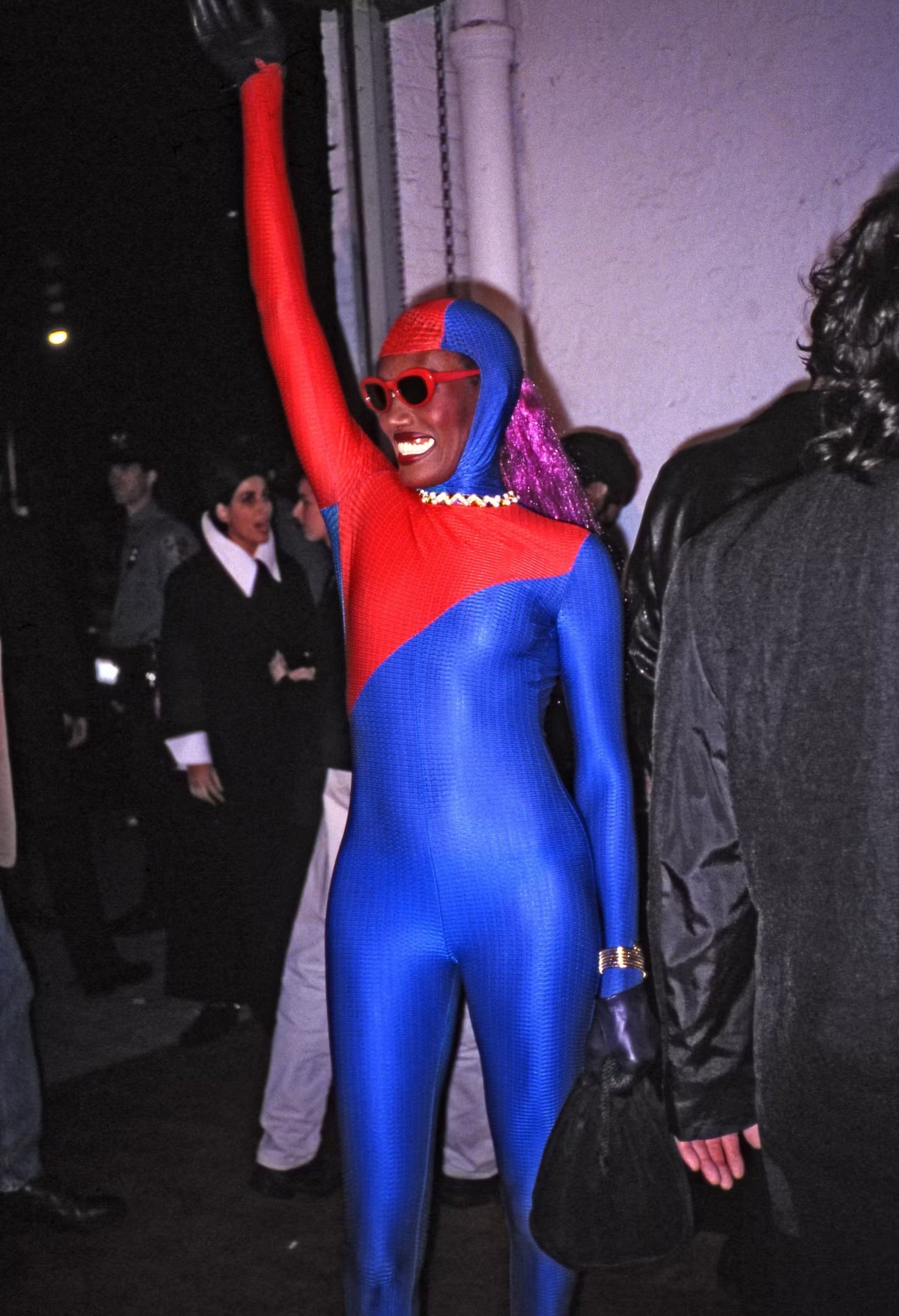 Grace Jones em roupa azul e vermelha brilhante segurando o braço no ar, usando óculos escuros e peruca roxa.