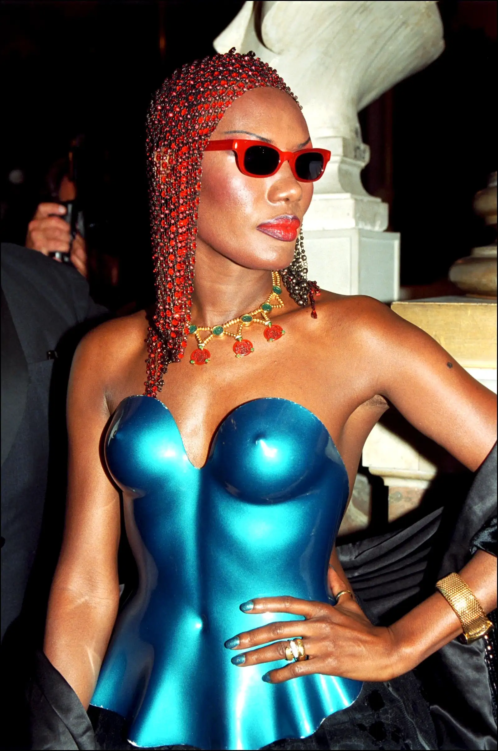 Grace Jones com lentes vermelhas, cabelo adornado e corpete azul cintilante.