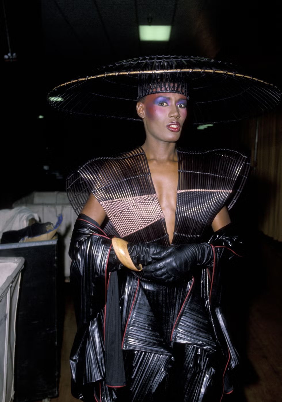 Grace Jones em traje futurista, com chapéu largo e maquiagem vibrante, posando diante de cortinas vermelhas.