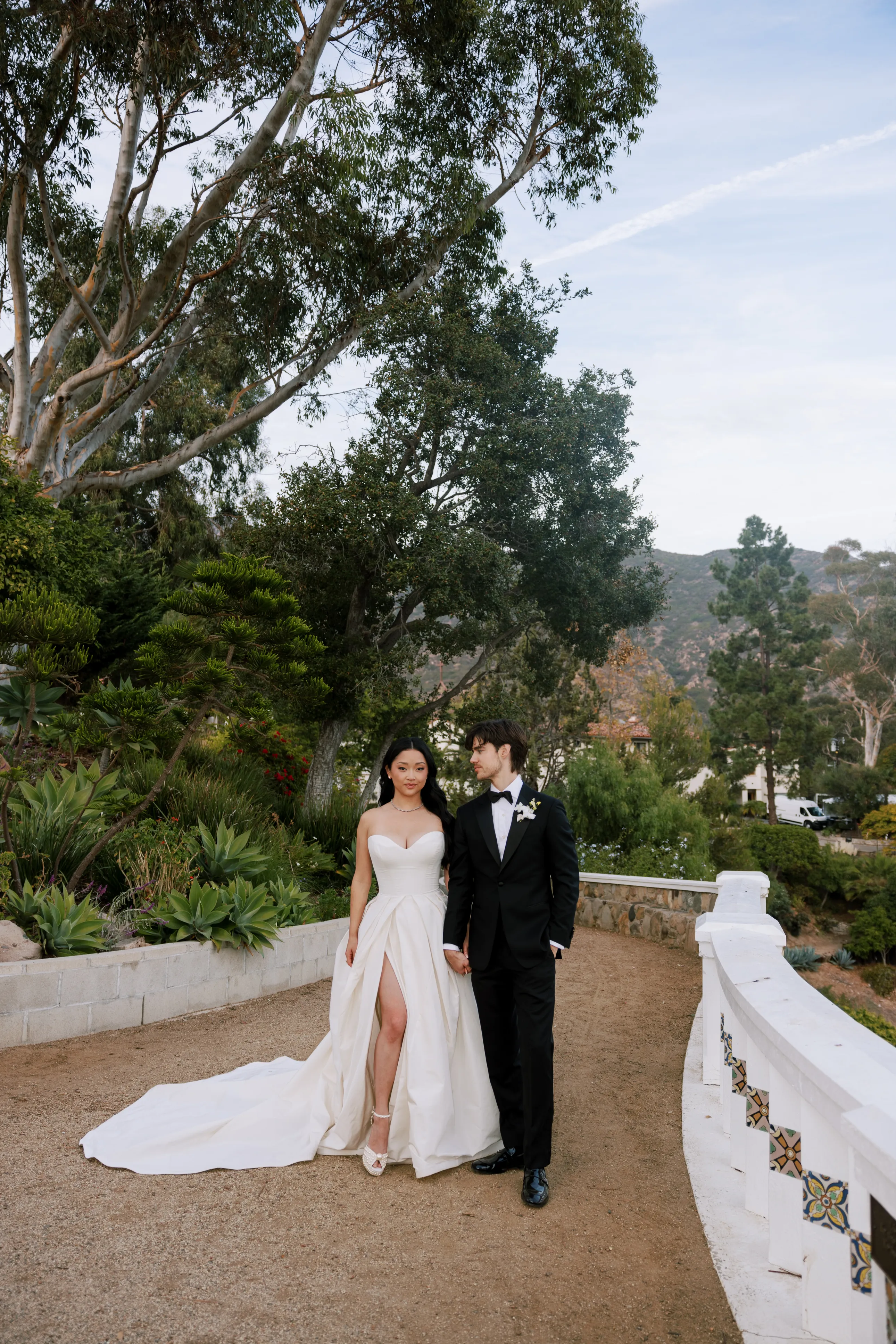 Um casal caminhando em um jardim, a noiva em vestido elegante com fenda e o noivo de terno preto. Vestidos de noiva das celebridades.