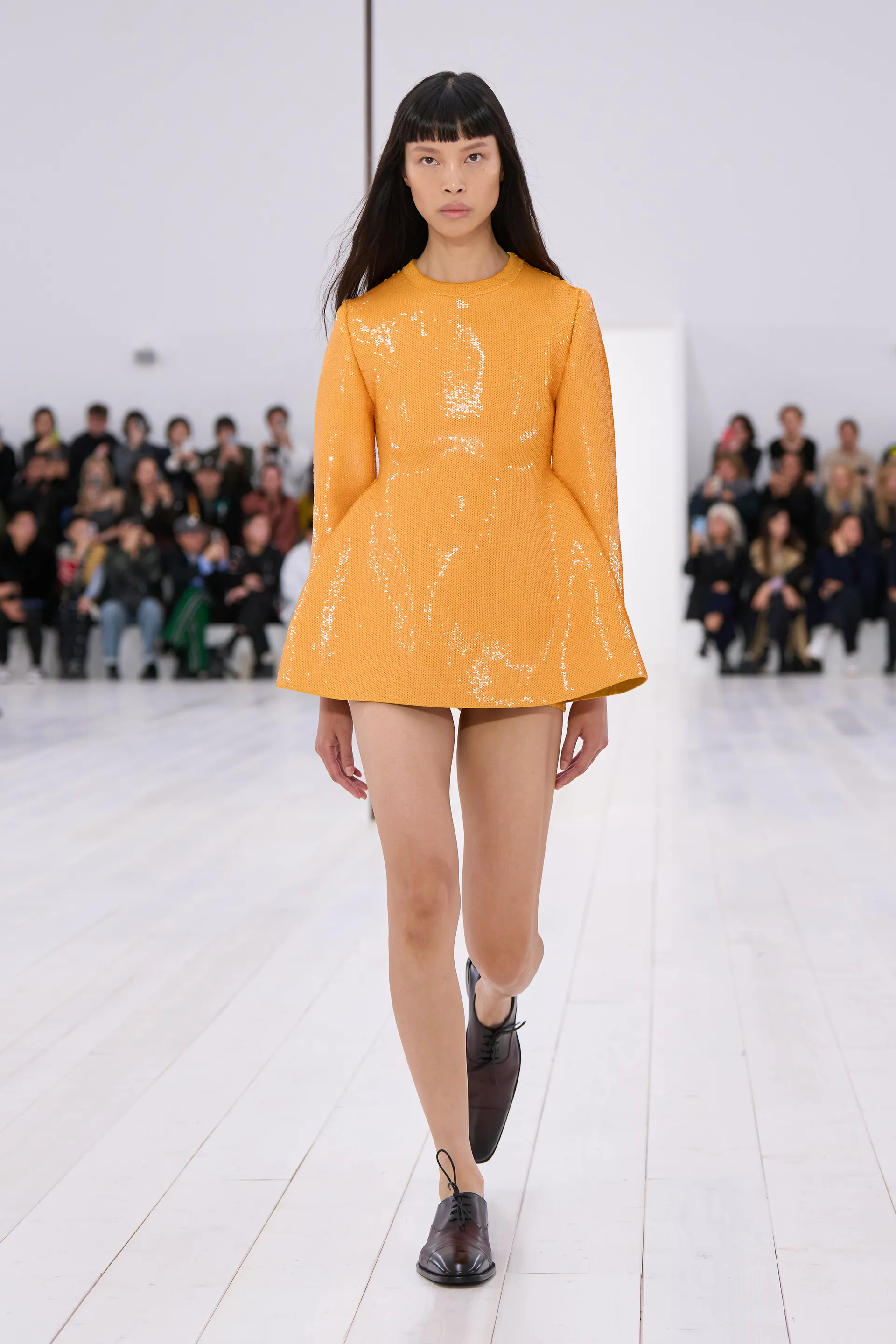 Modelo desfila com vestido laranja brilhante, símbolo das cores do verão 2025, em passarela com público ao fundo.