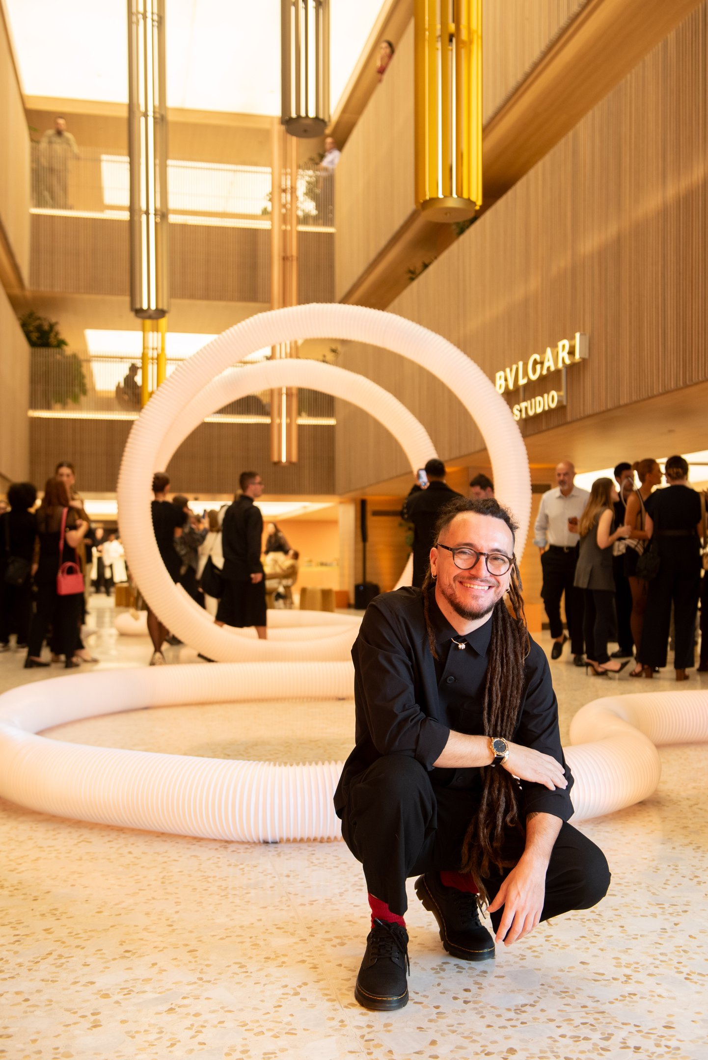 Ambiente sofisticado com homem sorridente em frente à instalação artística no Bvlgari Studio. Público interage ao fundo.