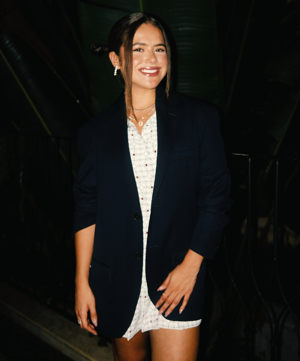 Mulher com blazer preto e vestido claro sorrindo em ambiente noturno, GP de São Paulo ao fundo.