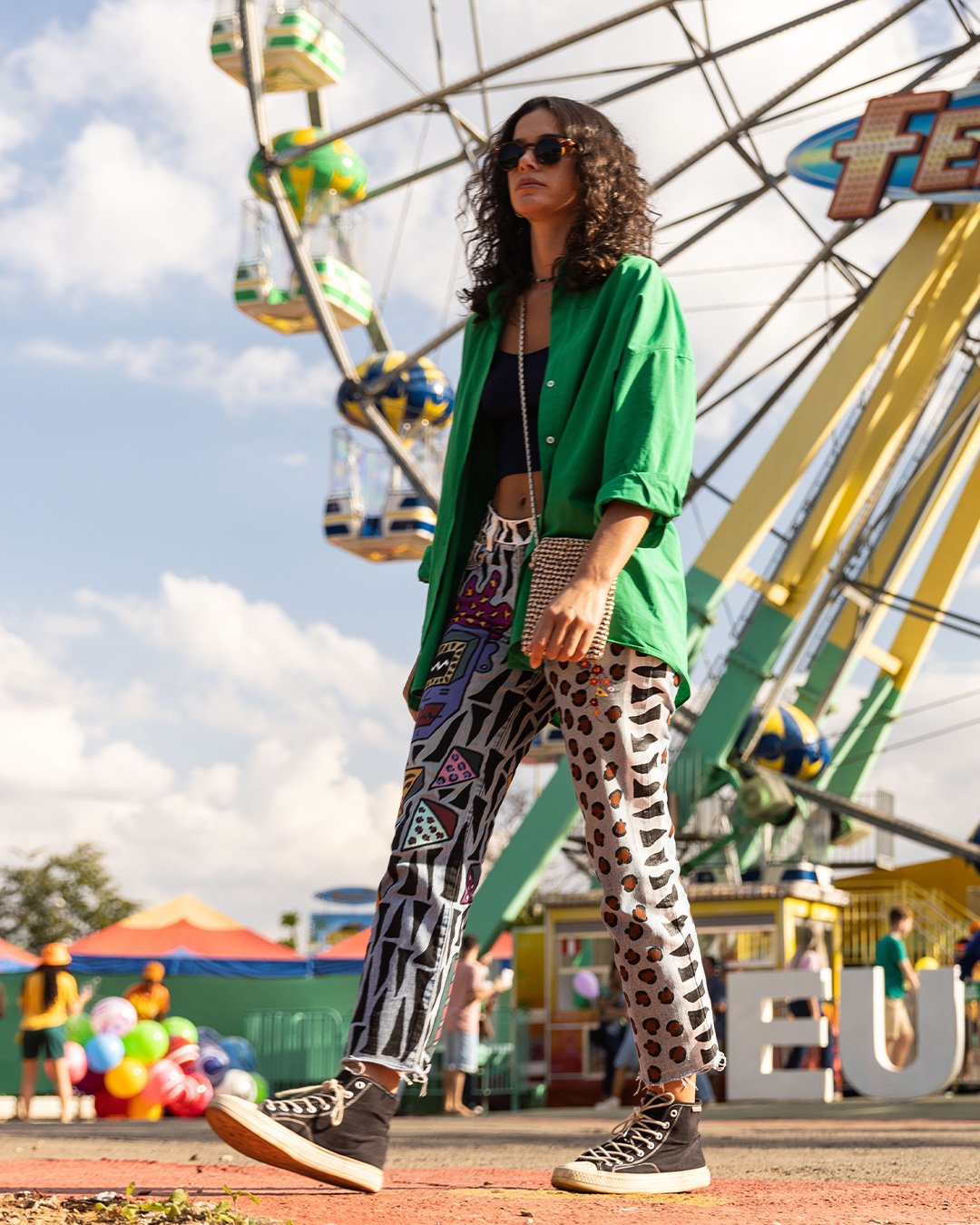 Mulher de óculos escuros e roupa colorida caminha em parque de diversões, com roda-gigante ao fundo e céu claro.