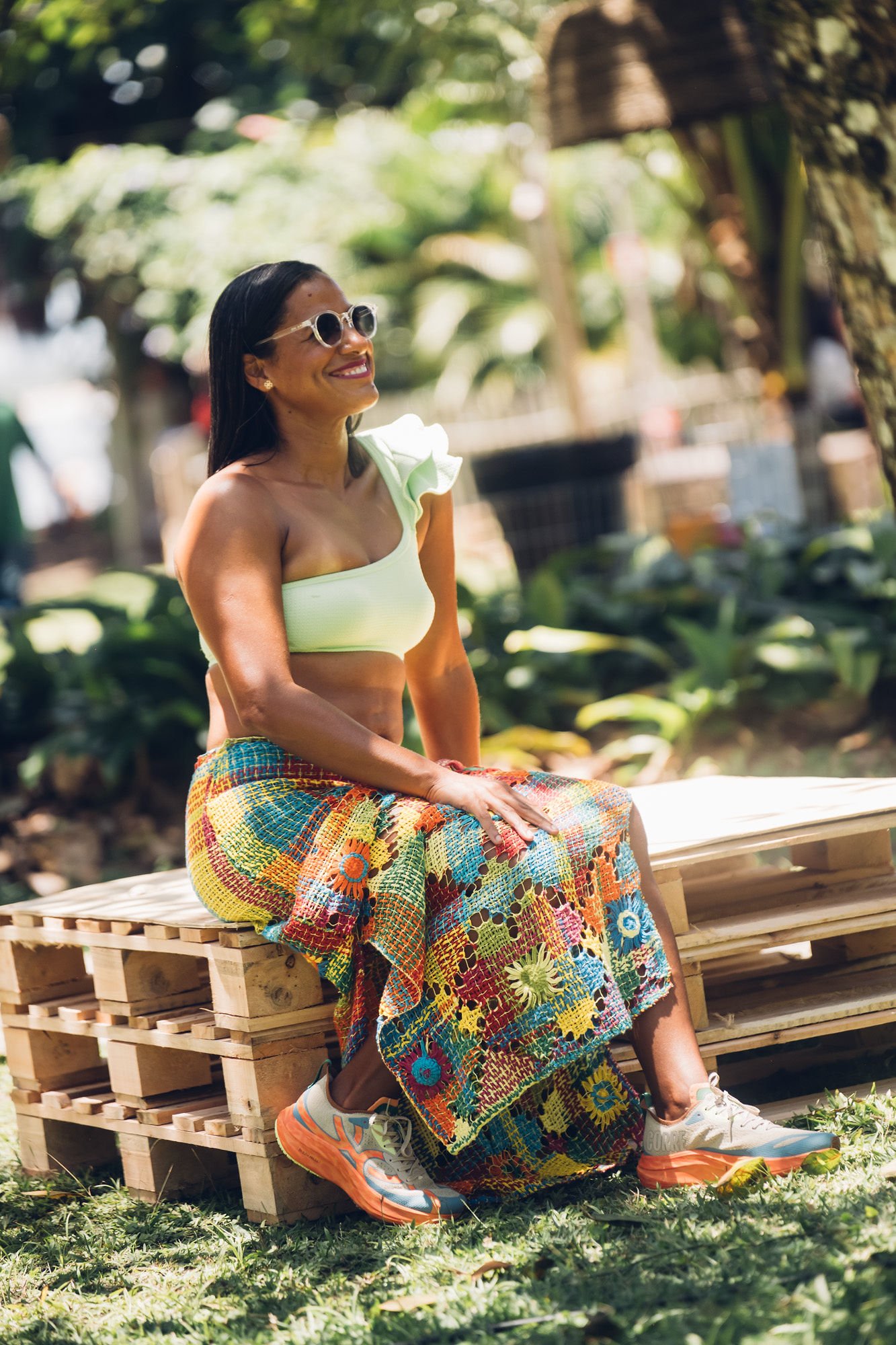 Pessoa sentada em banco de madeira, usando roupas coloridas e sorrindo ao ar livre. Os tênis têm detalhes chamativos.