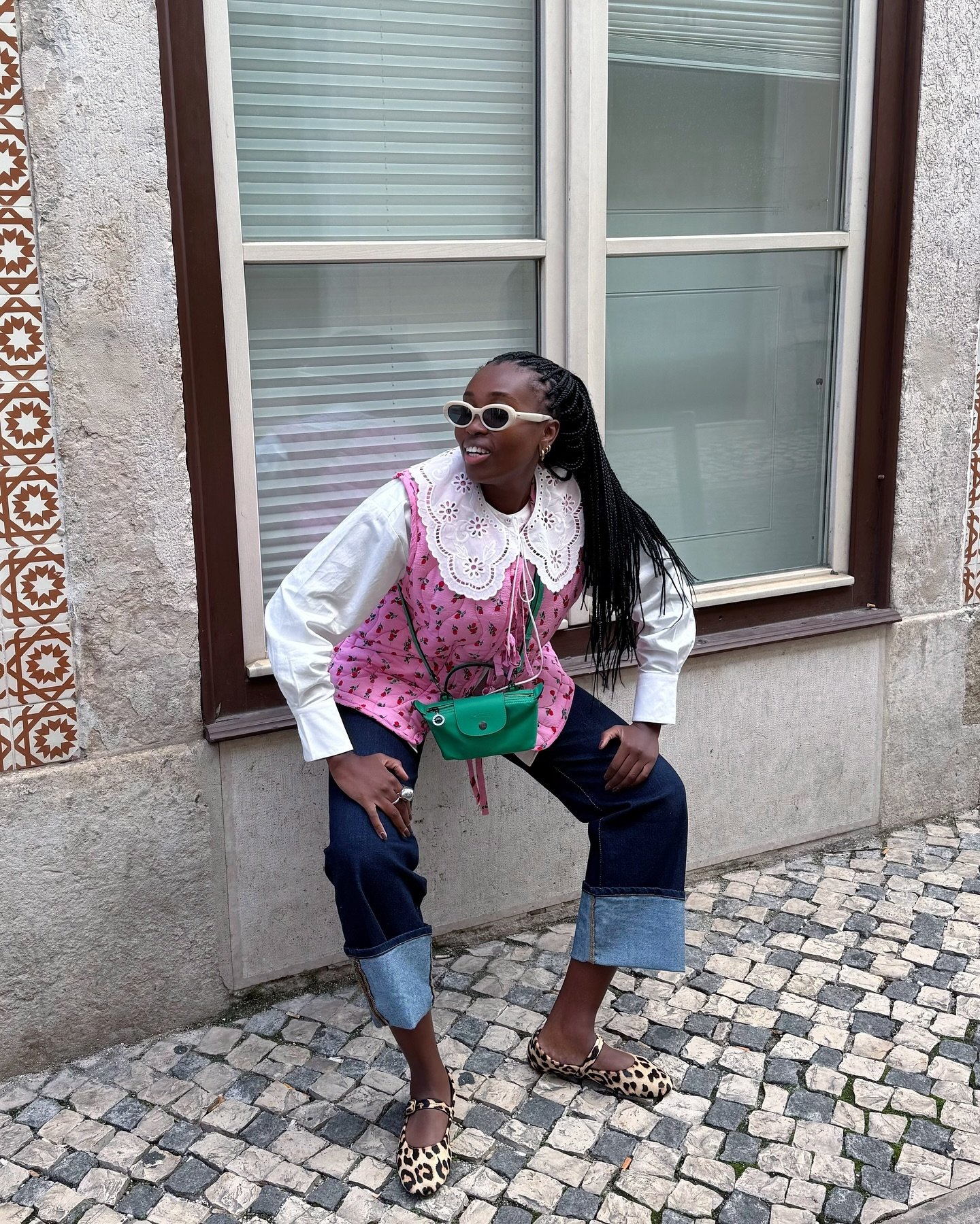 Pessoa sorridente com blusa rosa e gola Peter Pan branca, usando calça jeans dobrada e óculos de sol, em uma calçada de pedra.