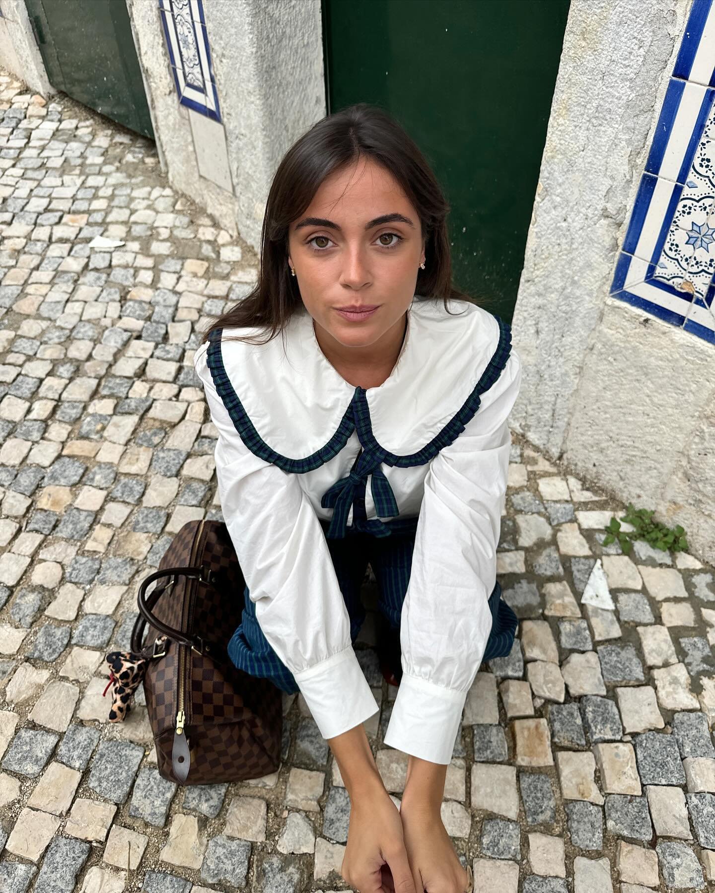 Mulher sentada em calçada de pedra, vestindo camisa branca com gola estilo Peter Pan. Bolsa marrom ao lado.