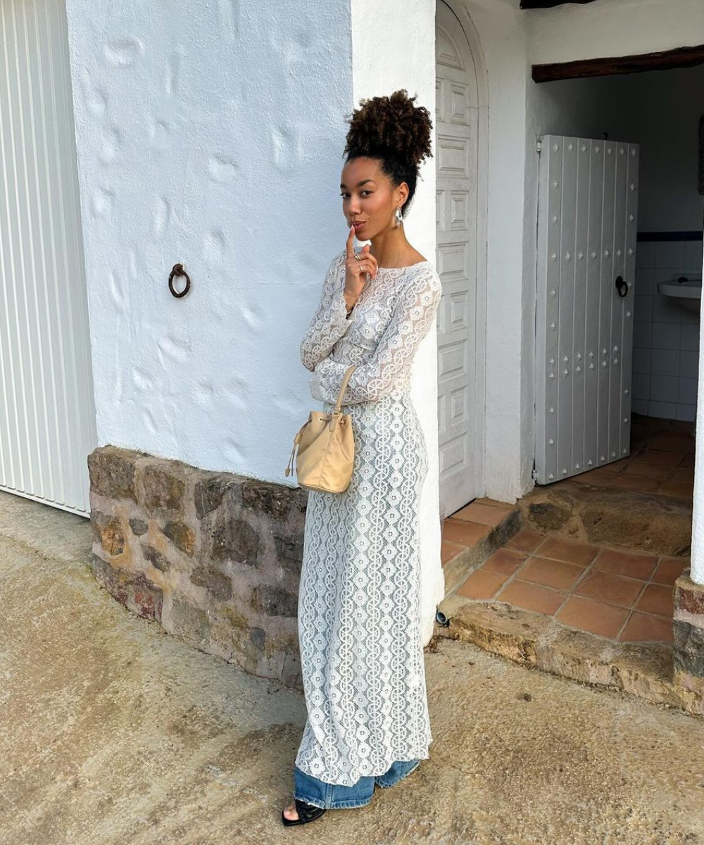 Mulher em vestido rendado branco sobre jeans, segurando uma bolsa bege, em frente a uma parede branca. Looks com jeans.