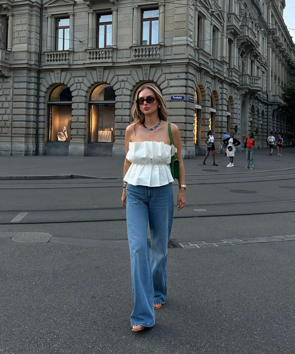 Mulher com blusa branca tomara-que-caia e jeans, caminhando em rua urbana. Elegante para looks de fim de ano.