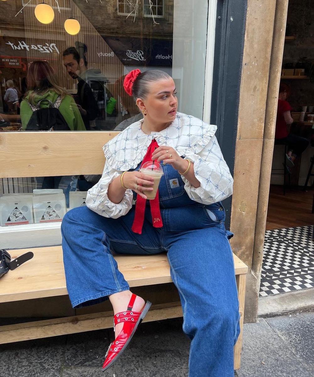 Mulher em jardineira jeans e blusa de babados, segurando café. Seu visual casual e moderno é ideal para looks de fim de ano.