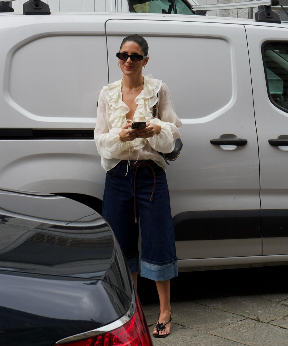 Mulher com blusa branca de babados e bermuda jeans escura. Acessórios: óculos de sol e sandálias pretas. Looks para o final do ano.