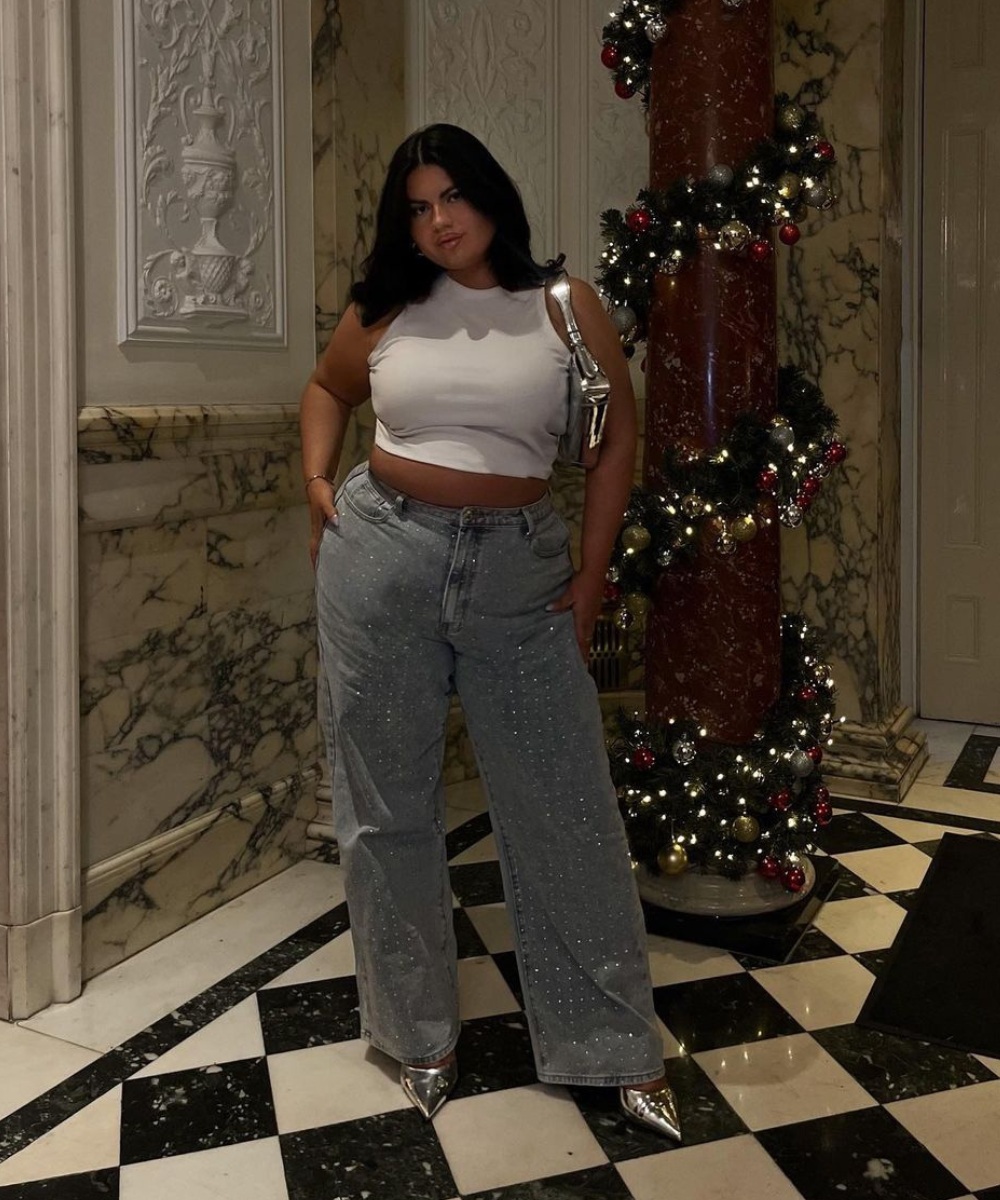 Mulher com top branco e jeans estilosos ao lado de coluna decorada para o Natal, look para o final do ano.