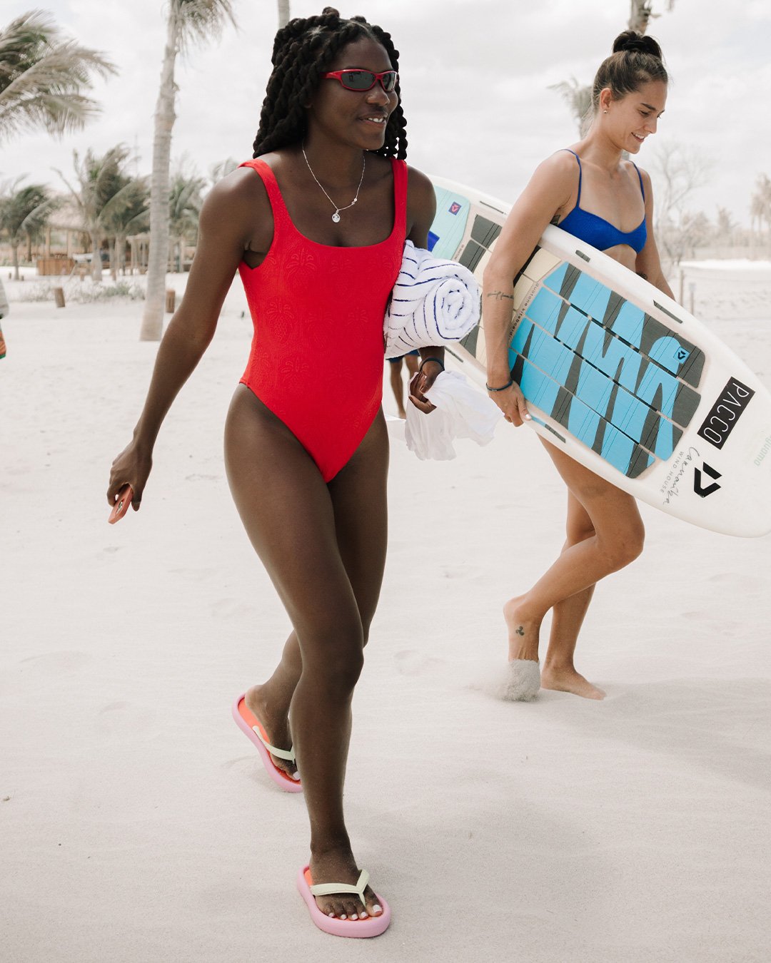 Mulher de maiô vermelho caminha na praia usando chinelo. Outra mulher segura prancha.