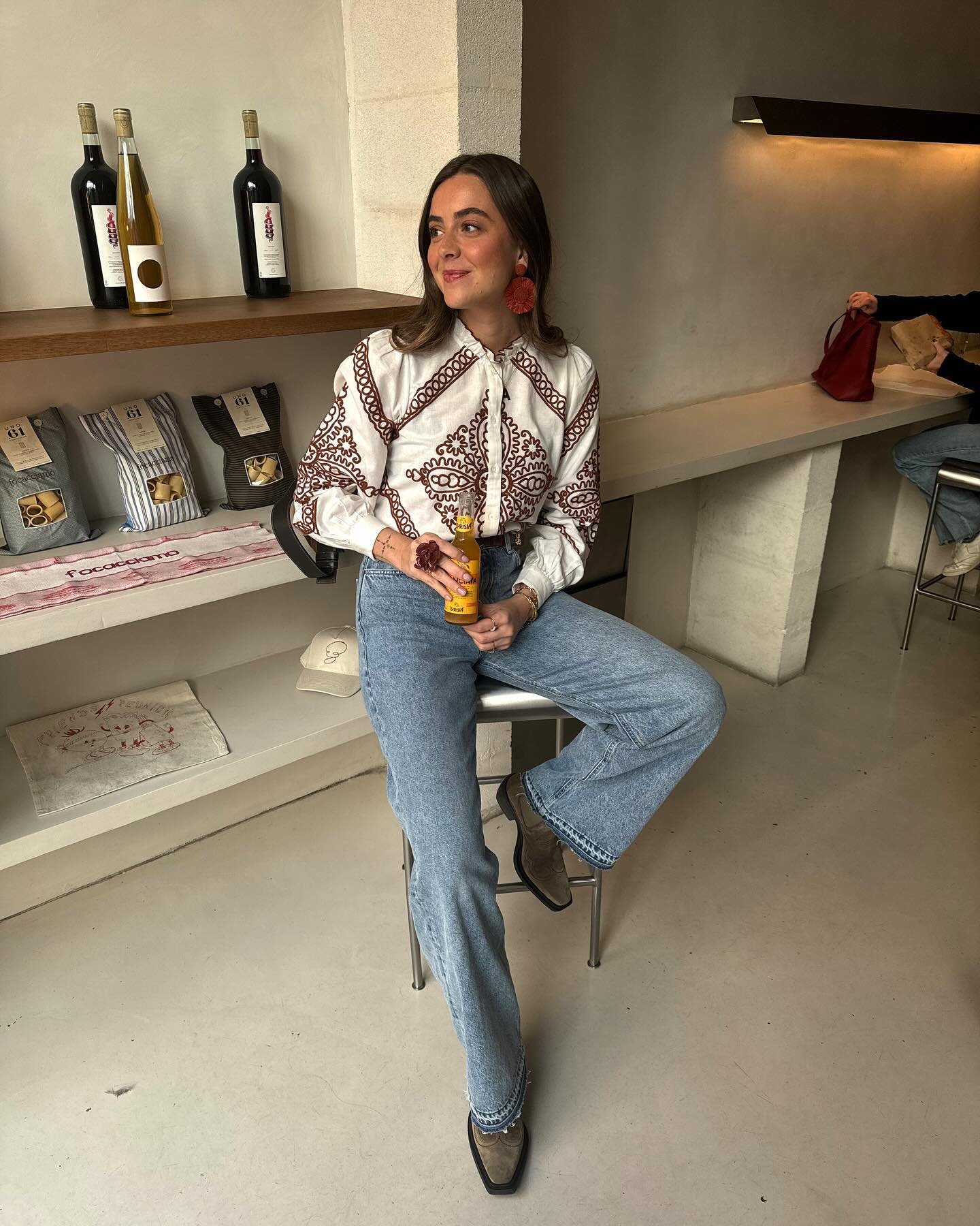 Mulher com camisa estampada e jeans, sentada em um café, segura uma bebida.éria looks elegantes de verão.