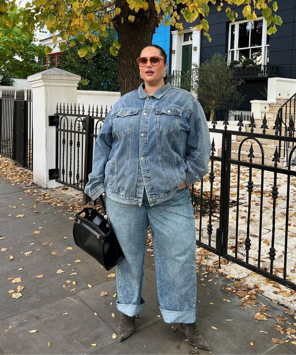 Mulher vestindo conjunto jeans com jaqueta e calça largas, segurando bolsa preta, em frente a uma casa. Look casual e bonito.