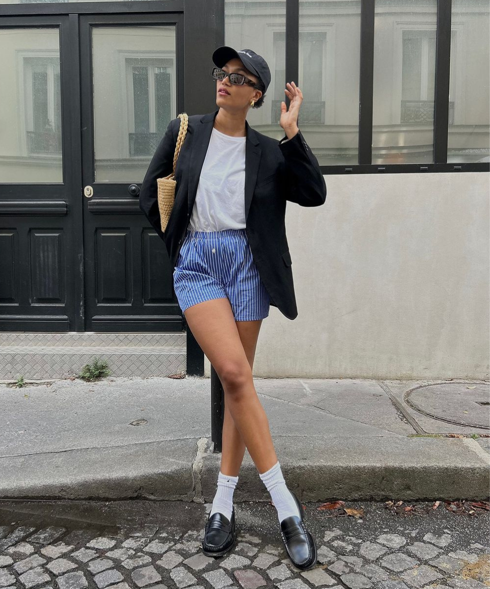 Pessoa com blazer preto, camiseta branca e shorts listrado azul em calçada urbana. Looks fáceis e bonitos.