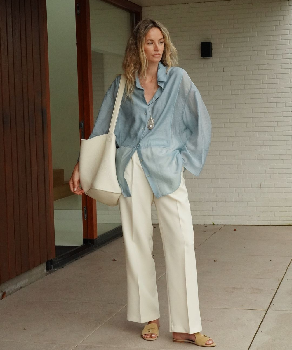 A pessoa está usando uma camisa azul clara, calça branca e sandálias, com uma bolsa grande. Looks fáceis e bonitos.