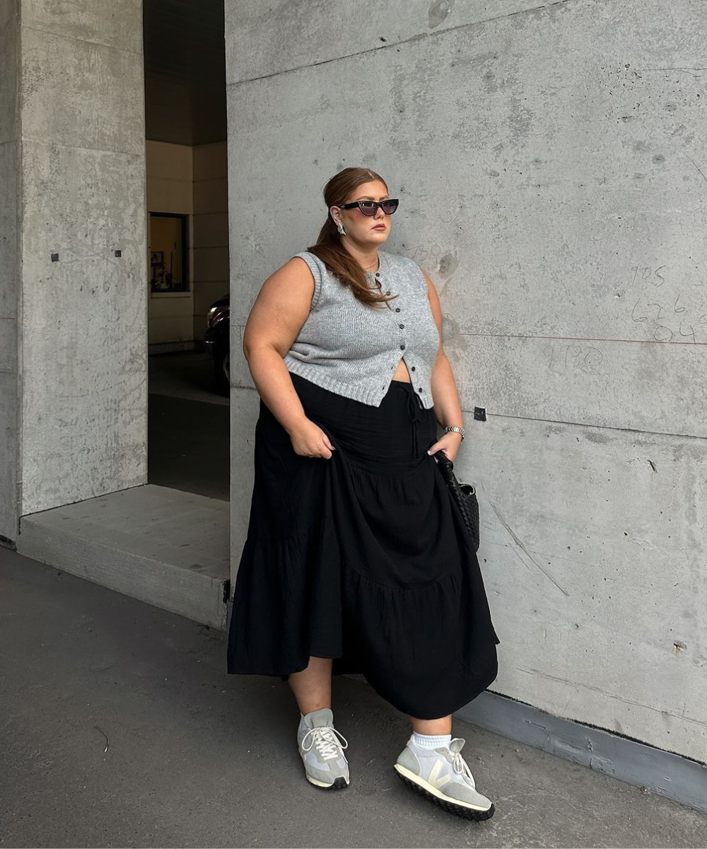 Mulher com colete cinza, saia preta longa e tênis cinza. Visual simples e estiloso em frente a um fundo de concreto.