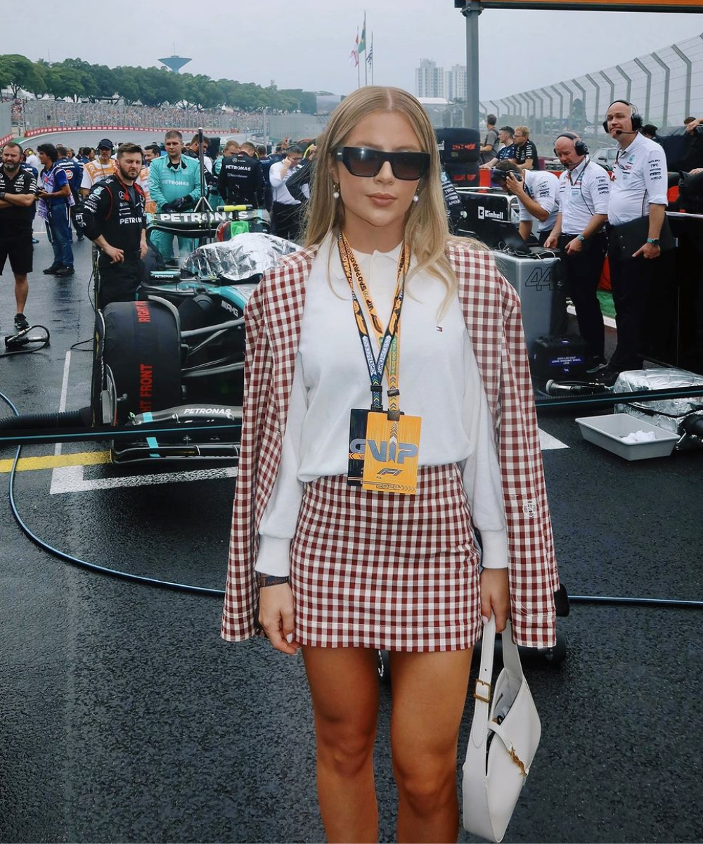 Mulher com roupa xadrez e crachá VIP ao lado de carro de corrida e membros da equipe. Atmosfera do GP de São Paulo 2024.