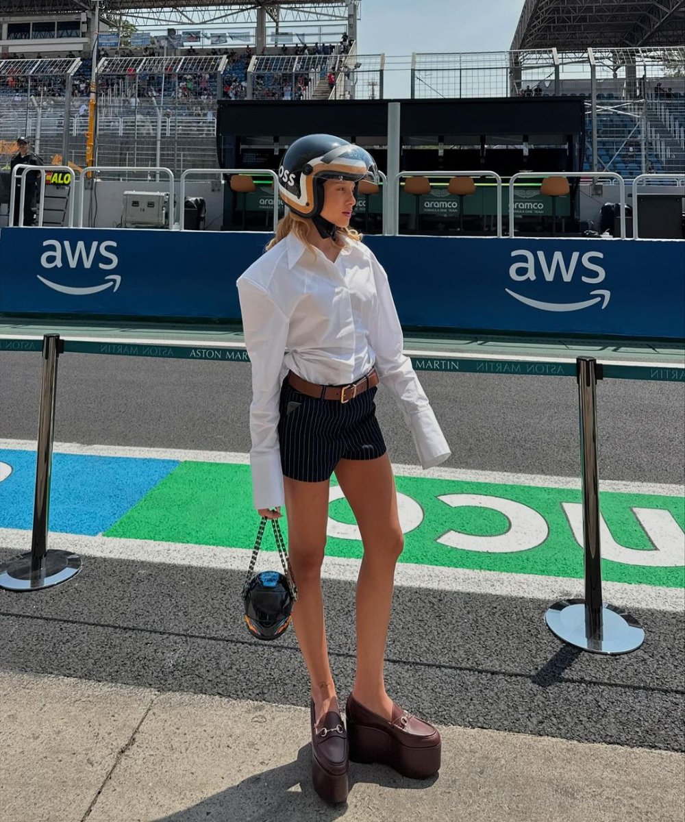 Pessoa com capacete no circuito de corrida do GP de São Paulo 2024, vestindo camisa branca, shorts listrados e sapatos altos.