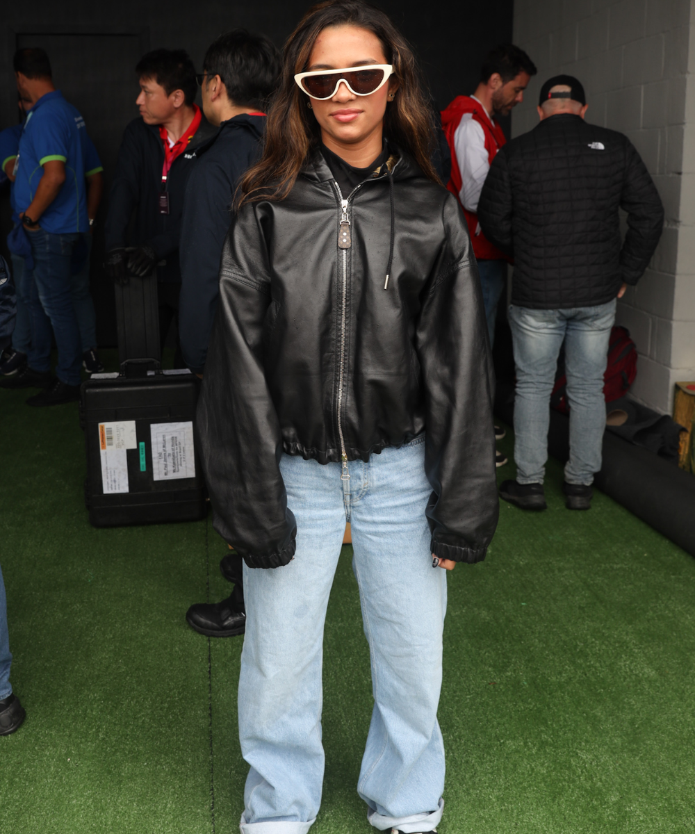 Pessoa com casaco de couro preto e óculos escuros posa em evento com várias pessoas ao fundo. Ambiente do GP de São Paulo 2024.