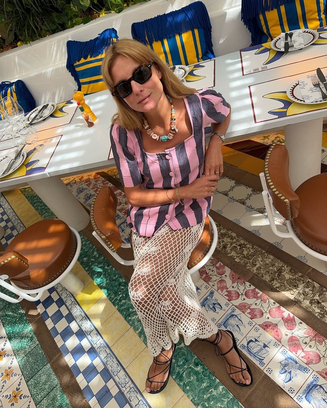 Mulher sentada em um restaurante, vestindo roupa de verão listrada e sandálias, com decoração de azulejos coloridos.