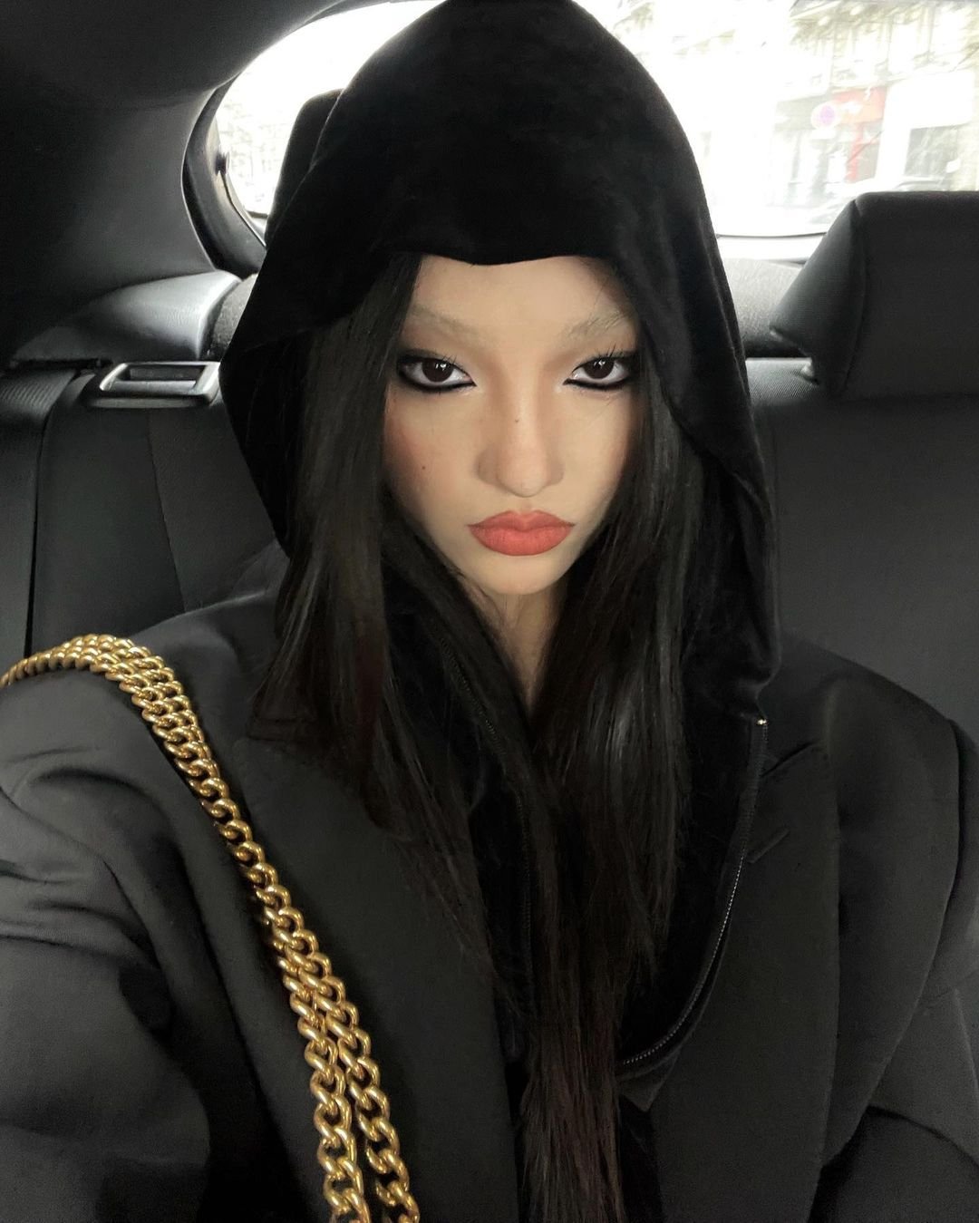 Mulher com capuz preto, maquiagem escura e expressão séria em carro. Estilo goth girl casual com corrente dourada.