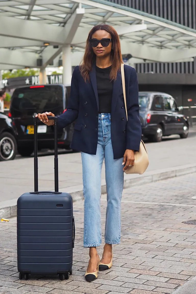 Mulher de pé segurando uma mala de mão para viagem em local urbano, vestindo blazer azul e jeans.