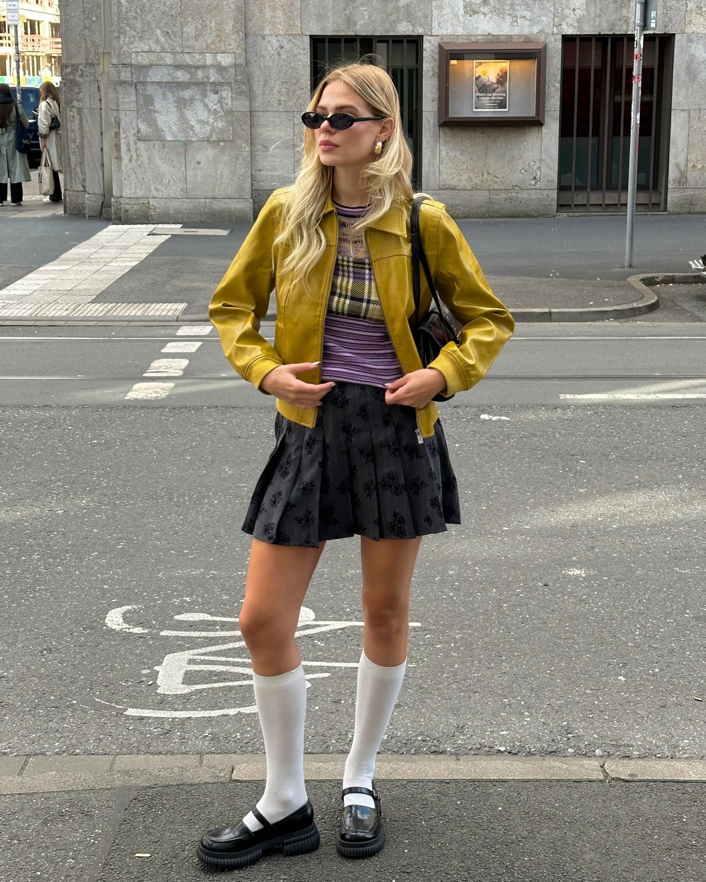 Mulher em roupa estilosa com casaco amarelo, saia preta e meias estilosas brancas, em uma rua urbana.