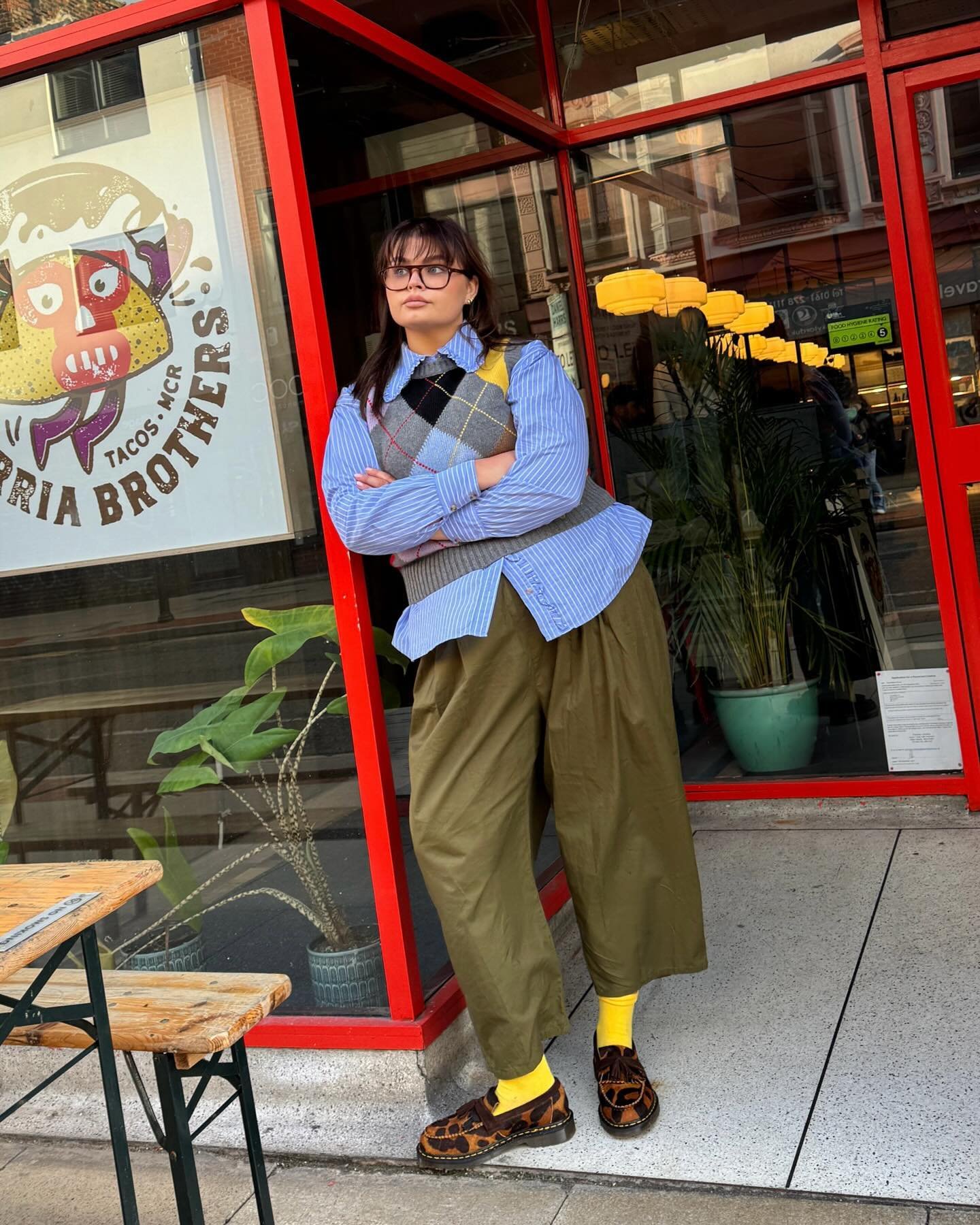 Pessoa de braços cruzados em frente a restaurante, usando camisa listrada, colete xadrez e meias estilosas amarelas.