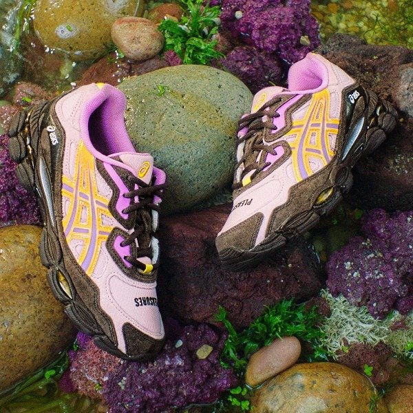 Tênis ASICS coloridos em um ambiente natural com pedras e vegetação ao redor.