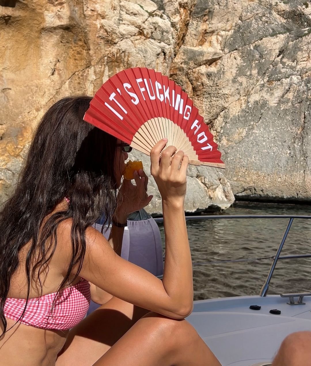 Mulher em biquíni usando leque com mensagem divertida em um barco. A cena sugere calor, destacando a importância de bons protetores solares.