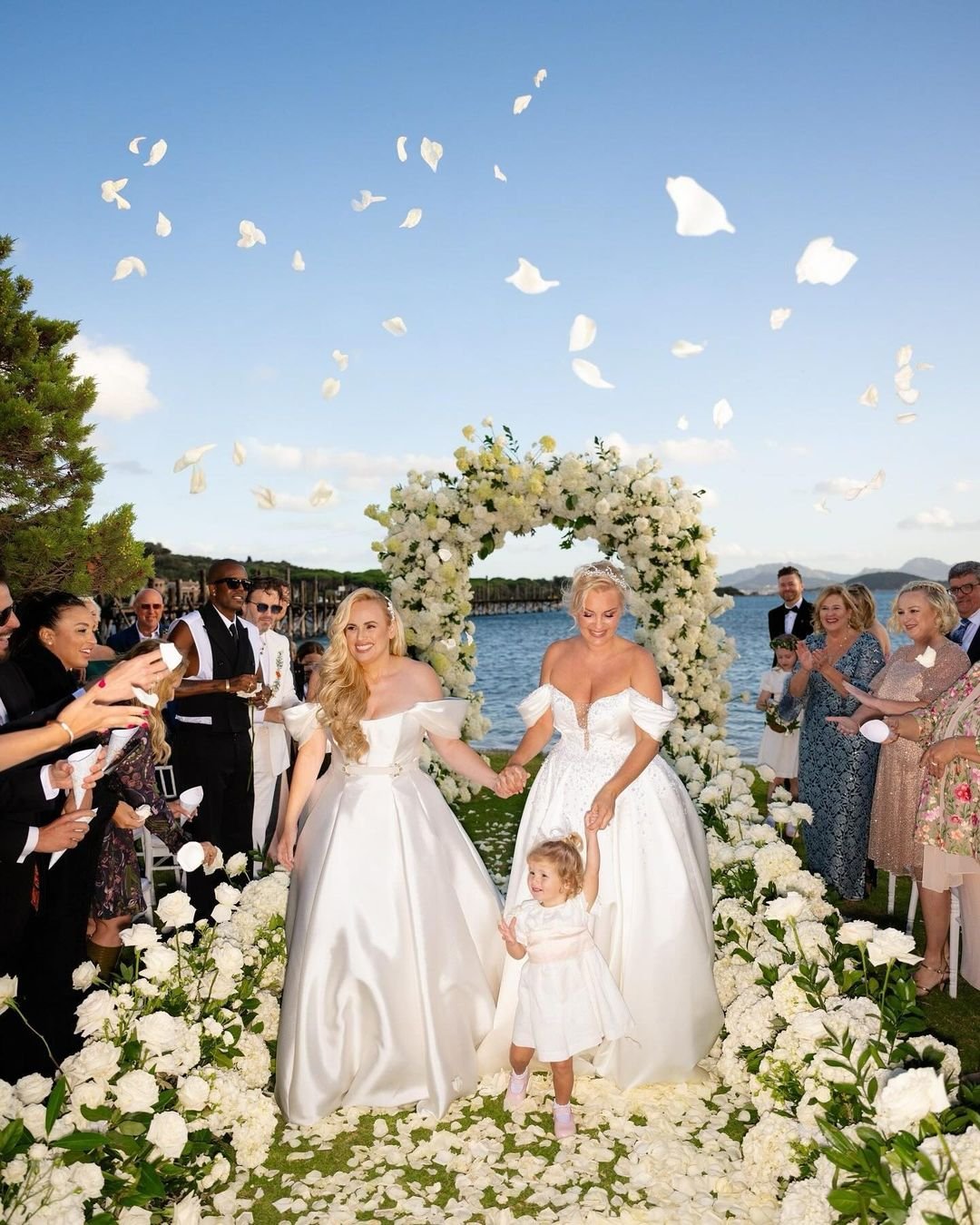 Duas noivas de braços dados em cerimônia, cercadas por pétalas e convidados sorrindo. Vestidos de noiva das celebridades.