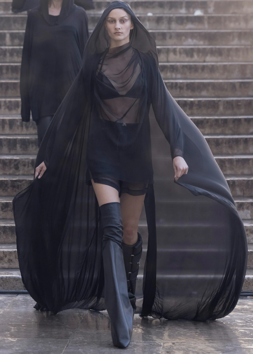 Desfile de moda com modelo usando vestido longo preto transparente, em estilo romance dark, com botas de cano alto.